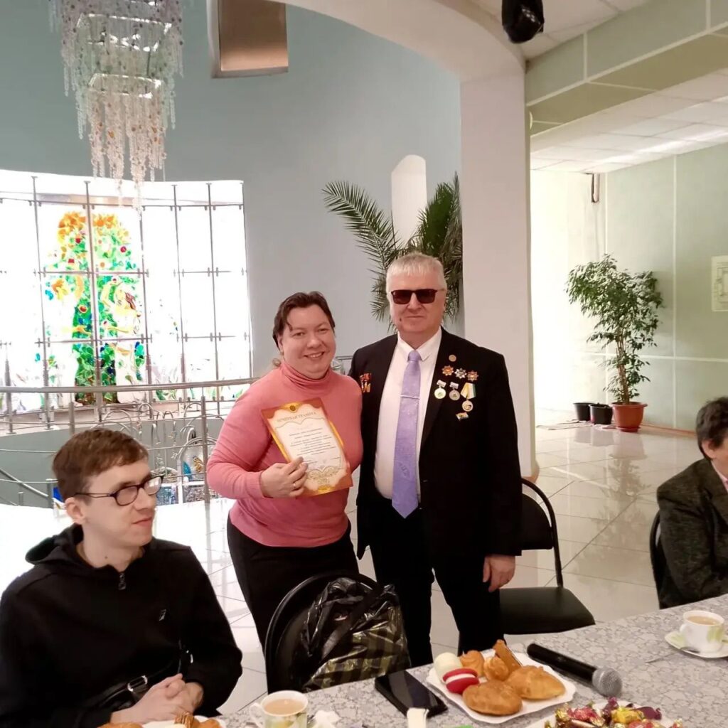 В центре женщина с грамотой и мужчина в костюме с медалями позируют в помещении; на столе чай, выпечка и микрофон, рядом сидят ещё люди.