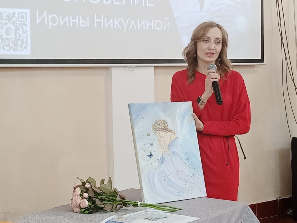 Женщина в красном платье с микрофоном стоит у проектора и показывает на мольберте картину в голубых тонах (девушка и бабочки). На столе перед ней букет розовых цветов и бумаги. На экране текст