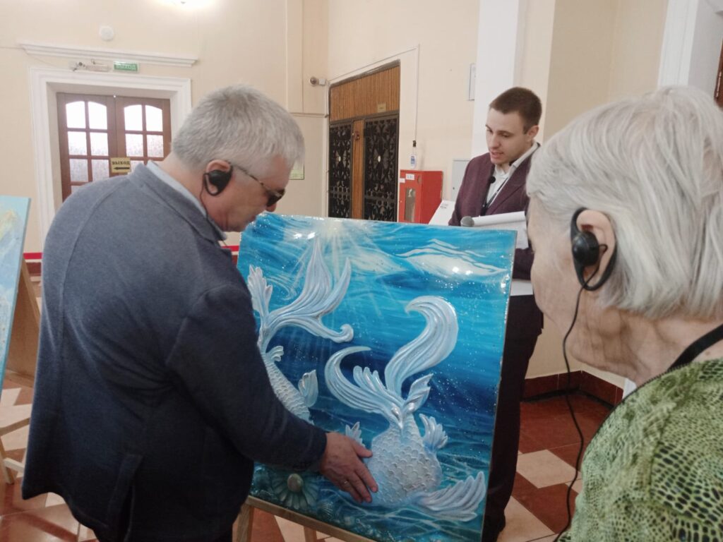 В помещении люди слушают аудиогид и рассматривают/ощупывают большую рельефную картину на мольберте: две бело-серебристые рыбы на ярко-синем “водном” фоне. 