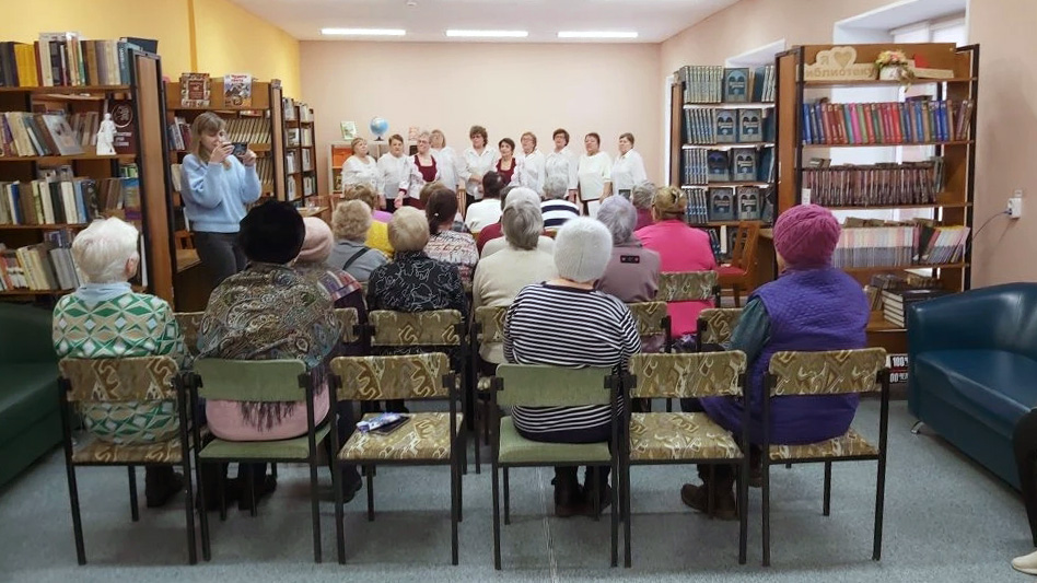 Библиотечный зал: сидит аудитория (в основном пожилые), впереди у стены выступает группа женщин в белых блузках, слева рядом стоит женщина в голубом свитере.
