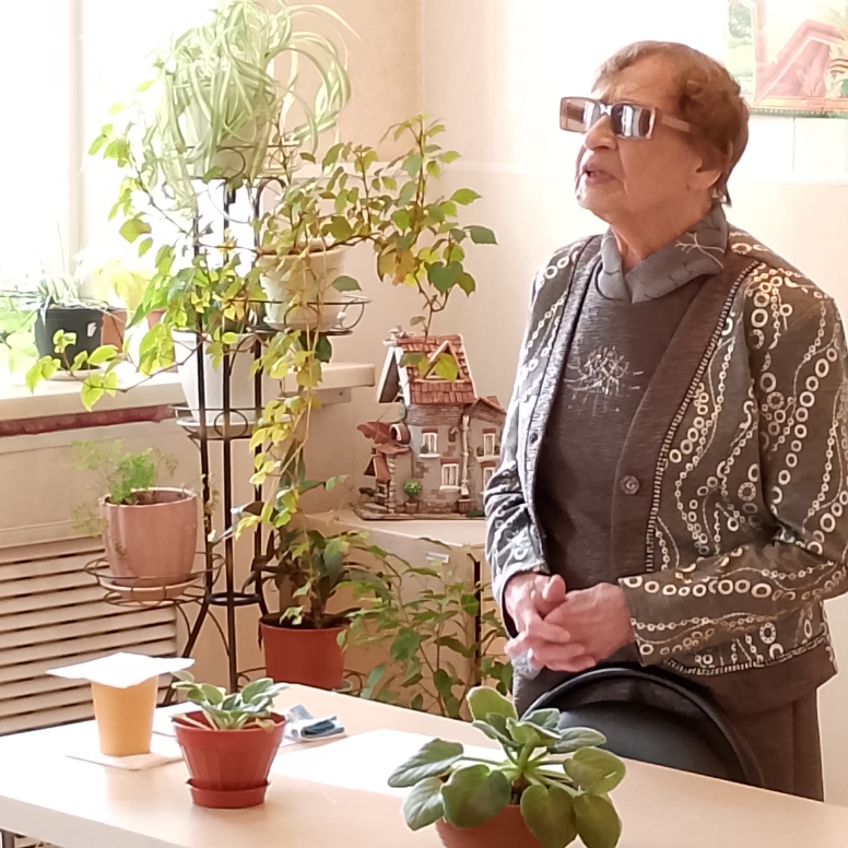 Пожилая женщина в очках стоит у стола с горшечными растениями; рядом окно и много растений на подставке.
