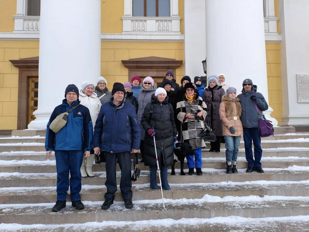 Около 20 человек в зимней одежде стоят на заснеженных ступенях перед зданием с большими белыми колоннами; в центре спереди женщина с белой тростью, рядом видна чёрная собака.
