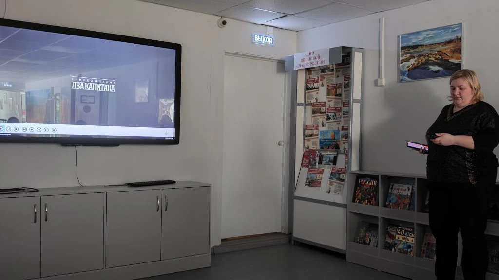 В помещении слева большой экран с видео и надписью «КИНОКОМПАНИЯ ДВАКАПИТАНА», по центру дверь со знаком «ВЫХОД», справа стенд «ДНИ РОДНОЙ СЛАВЫ РОССИИ», книжный стеллаж и женщина в чёрном со смартфоном.