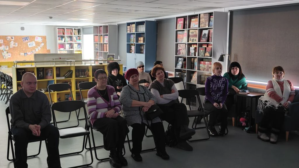 Зрители сидят на складных стульях в библиотечном помещении; на заднем плане книжные стеллажи, справа окна с серыми жалюзи.