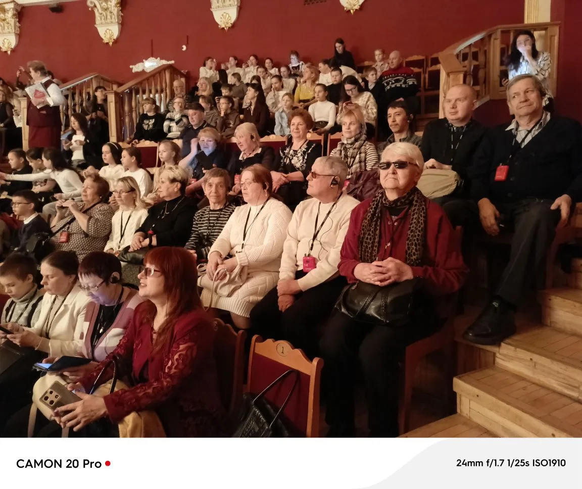 Зал с красными стенами и деревянными рядами, много зрителей разных возрастов, у многих бейджи; на переднем плане несколько пожилых людей, справа видна лестница со сидящими на ступенях людьми.