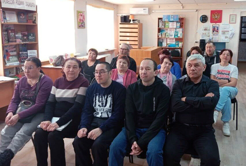 В комнате, похожей на библиотеку, несколько взрослых людей сидят рядами на стульях и смотрят в сторону камеры. На стенах книги, стенды, часы и герб; внизу