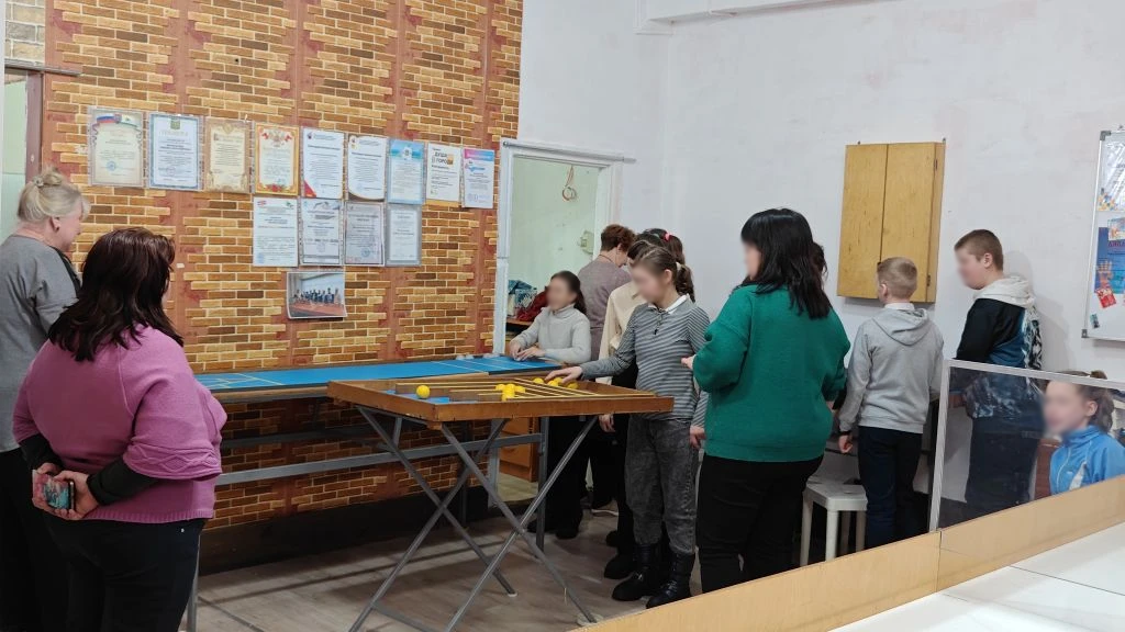 Группа взрослых и детей стоит в помещении вокруг длинного стола; на стене висят грамоты, рядом дверь и шкафчик.