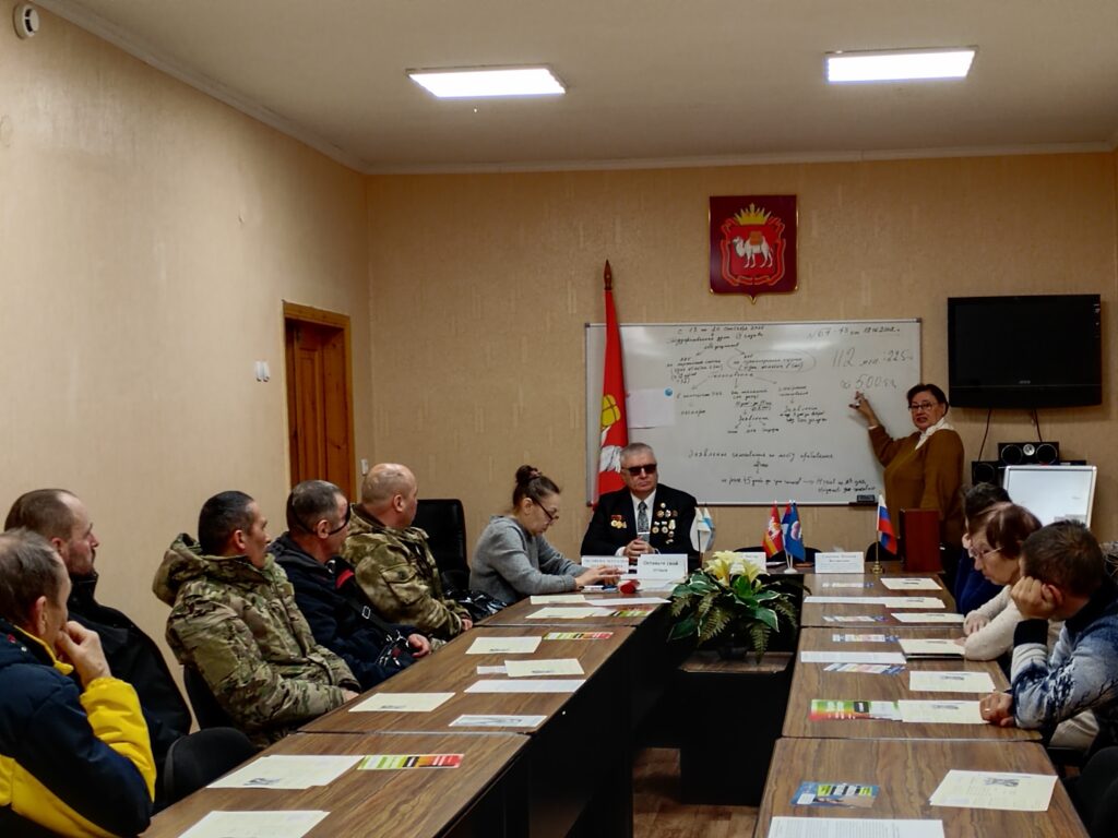 Совещание в зале: люди сидят за длинным столом, женщина у доски делает доклад; на столе документы и флажки, на стене герб, флаг и телевизор.