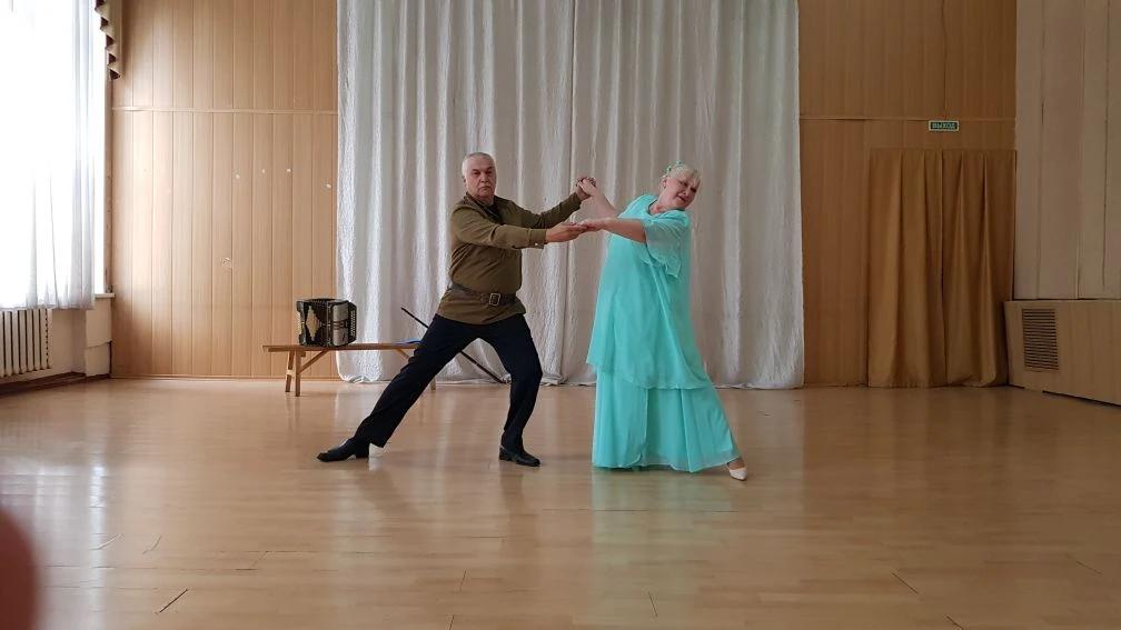 Пожилая пара танцует в зале: мужчина в коричневой рубашке и тёмных брюках держит за руки женщину в длинном бирюзовом платье на фоне белых занавесок.