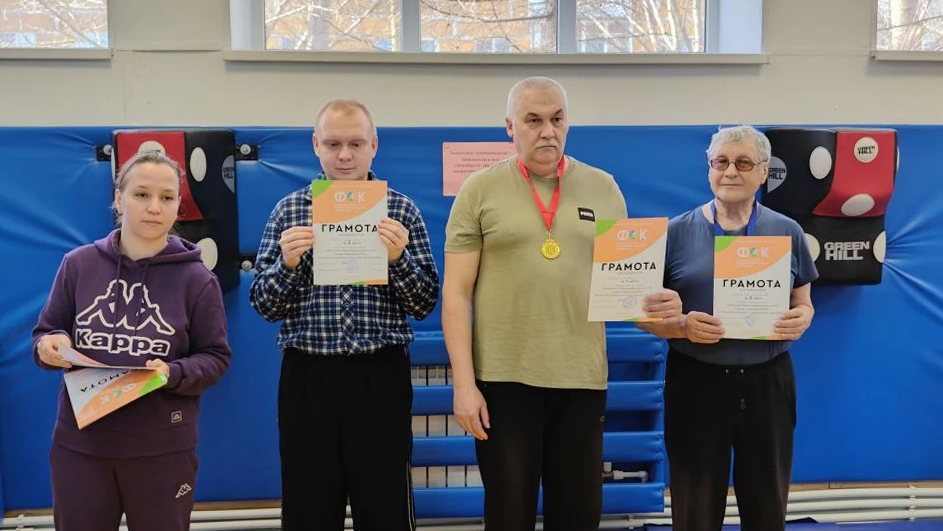 Четыре человека в спортзале стоят в ряд и держат грамоты; у мужчины в центре на шее медаль.