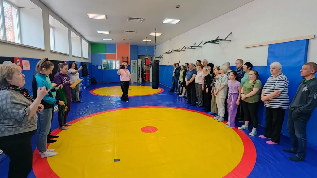 Спортзал с борцовским ковром. Люди стоят двумя рядами вдоль стен, в центре человек снимает на телефон. В глубине висят боксерские мешки, слева окна.