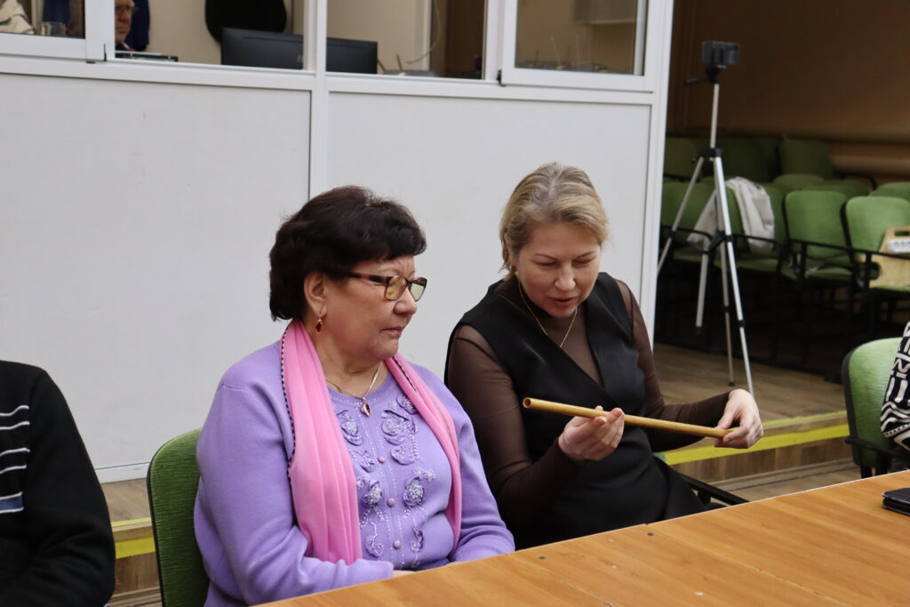 Две женщины сидят за столом; женщина справа держит и рассматривает длинную деревянную палочку. На заднем плане зелёные кресла, штатив и стеклянная перегородка.