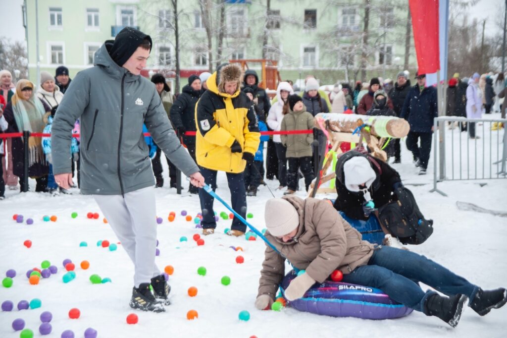 Зимнее мероприятие на улице: на снегу разбросаны разноцветные шарики, человек лежит на фиолетовой ватрушке SILAPRO, рядом двое людей, вокруг толпа за ограждением, идёт снег.