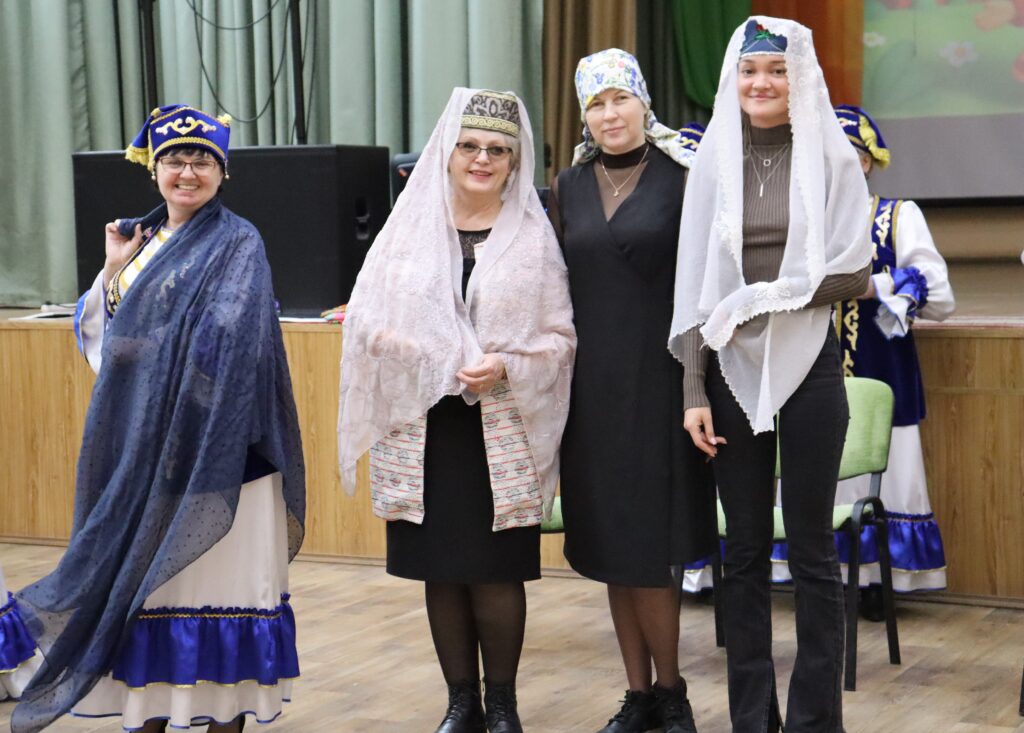 В помещении стоят четыре женщины и позируют; у нескольких на головах платки и тюбетейки/сценические костюмы. На заднем плане видны другие люди в похожих сине-белых костюмах и зелёный стул.