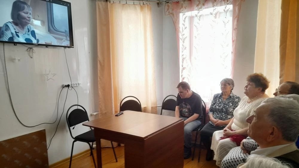 В комнате несколько человек сидят и смотрят телевизор на стене; в центре стол с пультом, справа яркое окно со шторами.