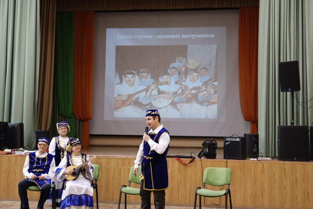 На сцене мужчина с микрофоном и несколько людей в сине-белых национальных костюмах; одна женщина держит щипковый инструмент. На экране слайд с фото детей-музыкантов и текстом «Группа струнно - щипковых инструментов».