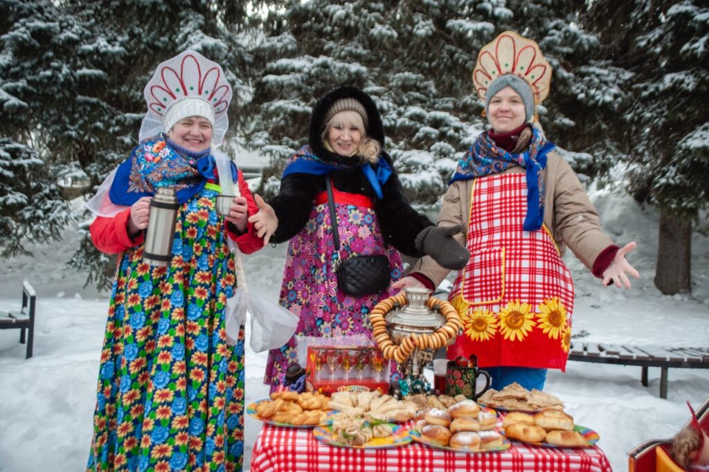Три женщины в народных зимних нарядах стоят на снегу у стола с самоваром, баранками и выпечкой на красно-белой клетчатой скатерти; на фоне заснеженные ели.