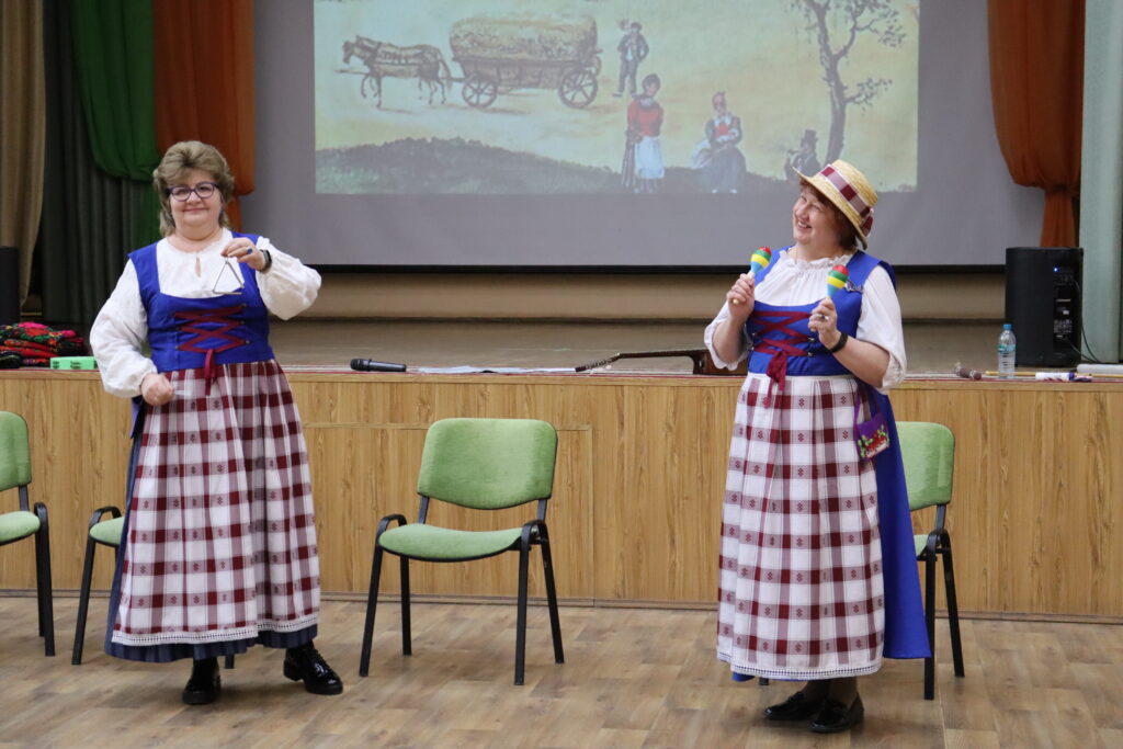 Две женщины на сцене в народных костюмах: слева держит музыкальный треугольник, справа в шляпе держит две маракасы и улыбается. На сцене зелёные стулья, сзади экран с рисунком повозки с лошадью.