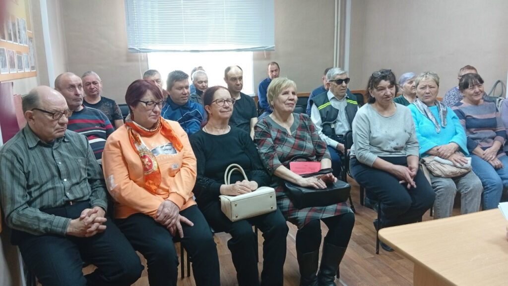 несколько рядов людей сидят на стульях и слушают. На заднем плане окно с жалюзи, слева на стене стенд с фотографиями.