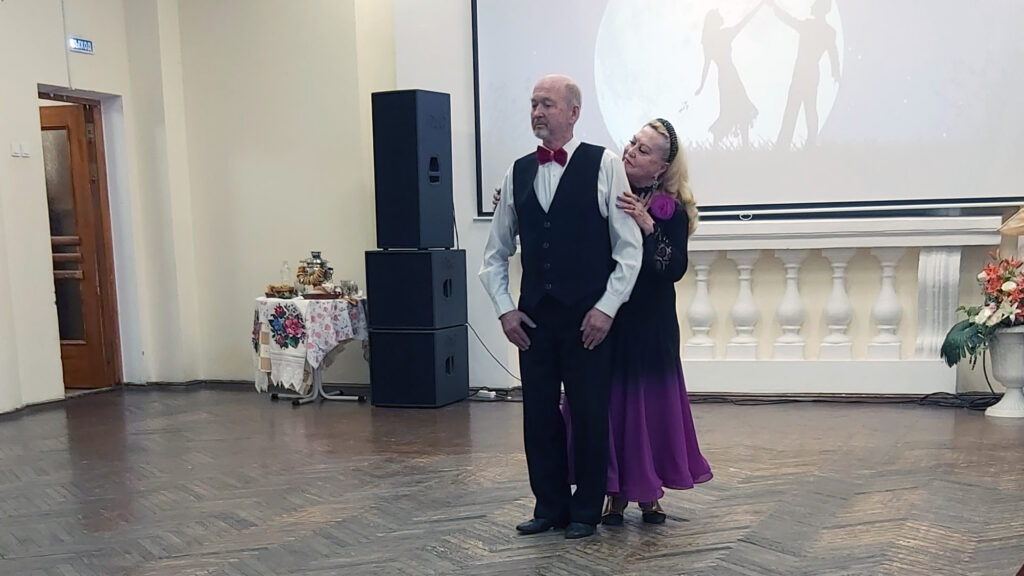 Мужчина в жилете и с красной бабочкой стоит на сцене, а женщина в длинном фиолетовом платье стоит позади и обнимает его за плечи. На заднем плане экран с силуэтом танцующей пары и большая колонка; слева стол с самоваром.