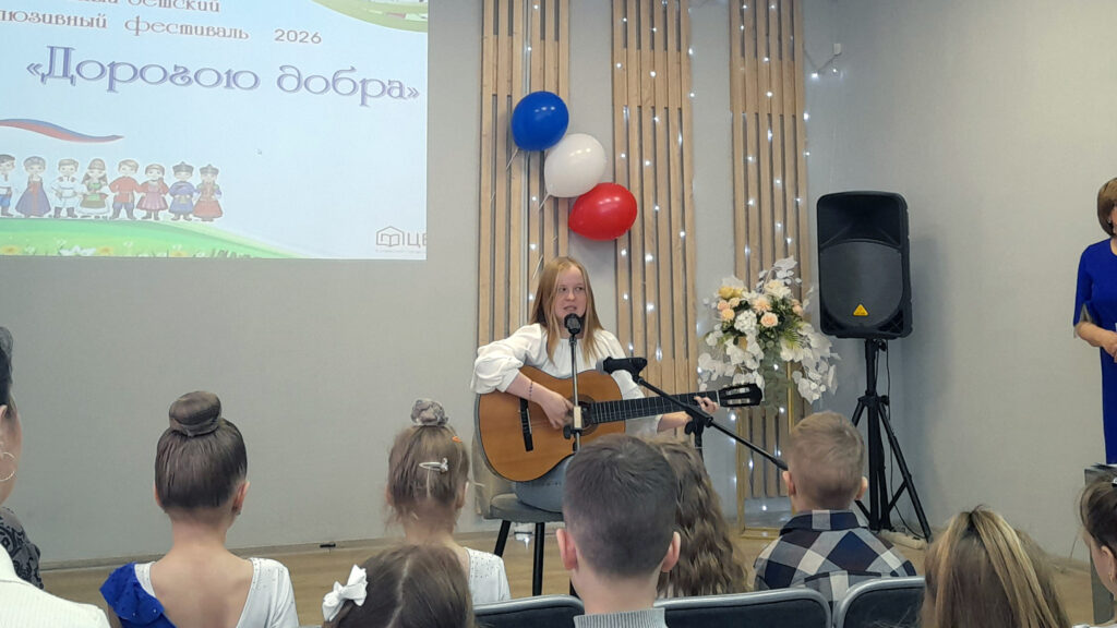 На сцене девочка со светлыми волосами сидит на стуле и играет на гитаре, поёт в микрофон. В зале перед ней сидят дети. 