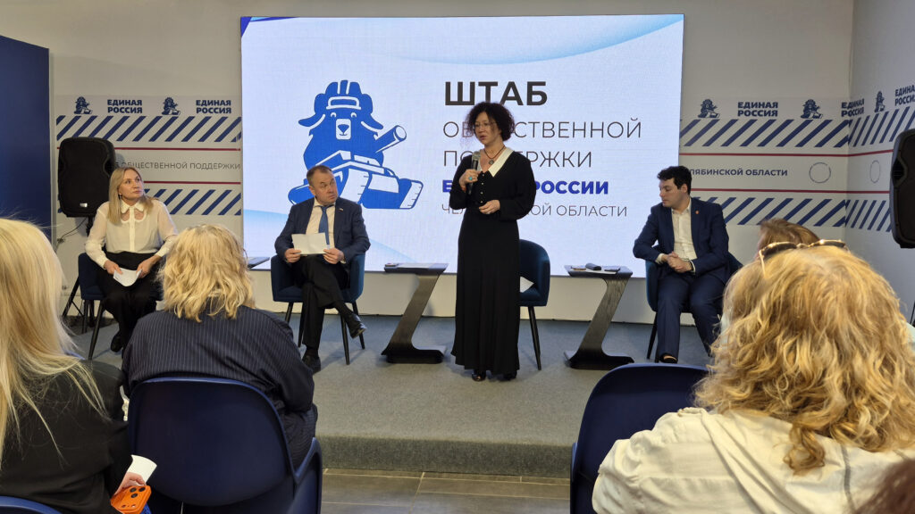 наталья лугачёва в чёрном платье стоит и говорит в микрофон, трое рядом сидят