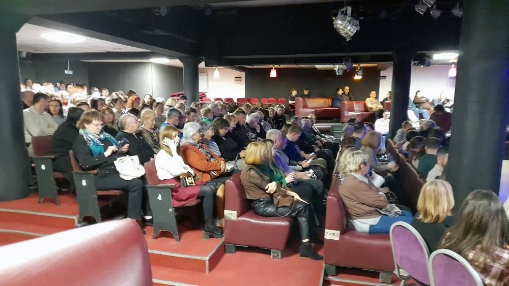 Зал со зрителями: много людей сидят рядами на бордовых креслах, слева видны ступени с номерами мест. Освещение приглушённое, на потолке прожекторы.