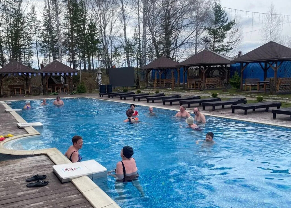 Открытый бассейн на улице, в воде купается группа взрослых людей, вокруг деревянный настил, лежаки и несколько беседок; на заднем плане деревья и пасмурное небо.