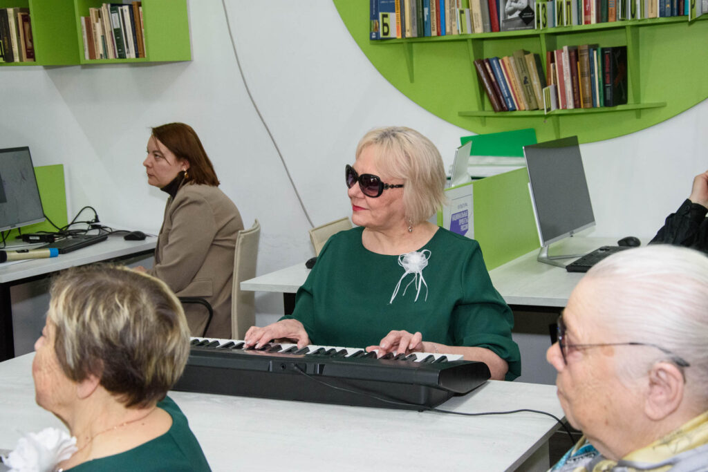 В помещении с компьютерами и книжными полками женщина со светлыми короткими волосами в тёмно-зелёном платье и тёмных очках играет на электронном клавишном инструменте на столе. Позади сидит женщина в бежевом пиджаке за компьютером; на переднем плане частично видны ещё два человека.