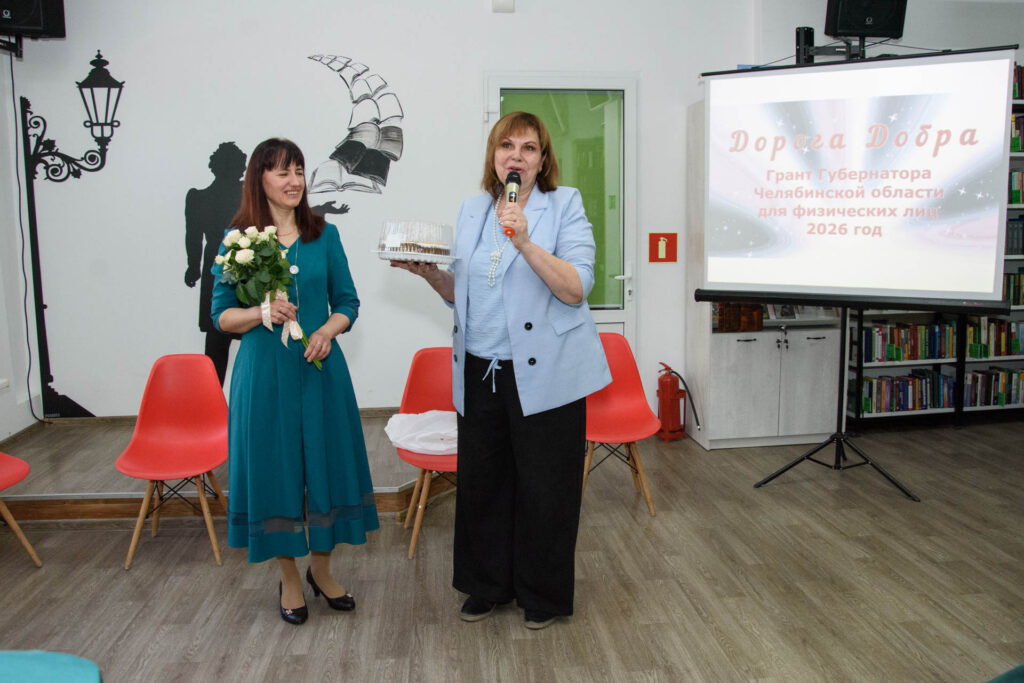 Две женщины на мероприятии: одна в голубом пиджаке говорит в микрофон и держит торт в контейнере, другая в бирюзовом платье держит букет белых цветов. 