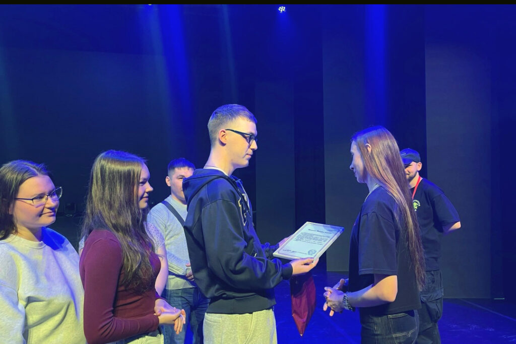На сцене под синим светом парень в очках вручает девушке с длинными волосами документ в рамке; рядом стоят ещё несколько людей в очереди.
