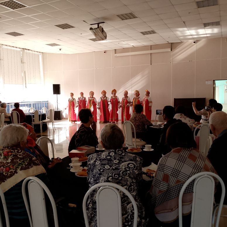 В зале люди сидят за столами, а впереди выступает женский ансамбль в красных народных костюмах.