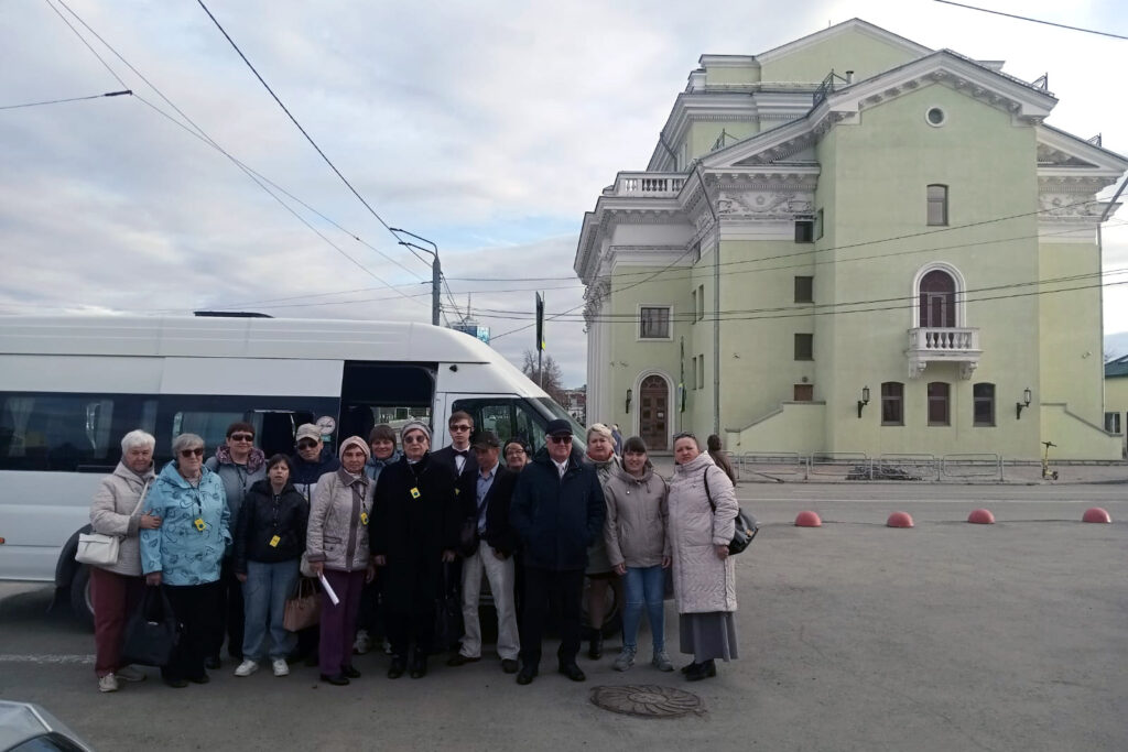Группа из примерно пятнадцати человек стоит на улице перед белым микроавтобусом.
Люди одеты в куртки и пальто, большинство среднего и пожилого возраста.
На заднем плане крупное светло‑зеленое здание зала органной и камерной музыки с колоннами и балконом, 