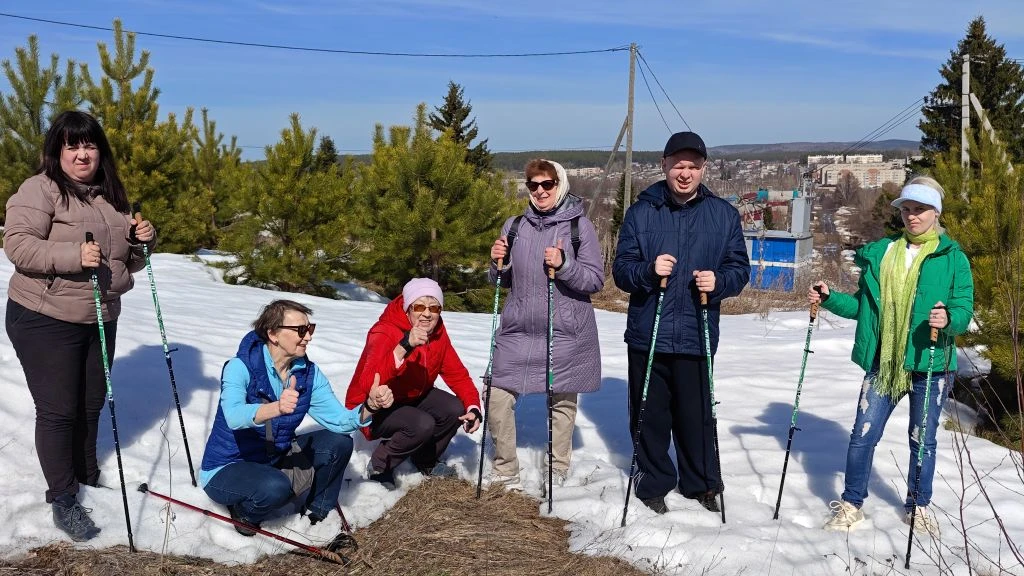 Шесть человек с палками для скандинавской ходьбы позируют на снегу у сосен; на заднем плане виден город и линии электропередачи.