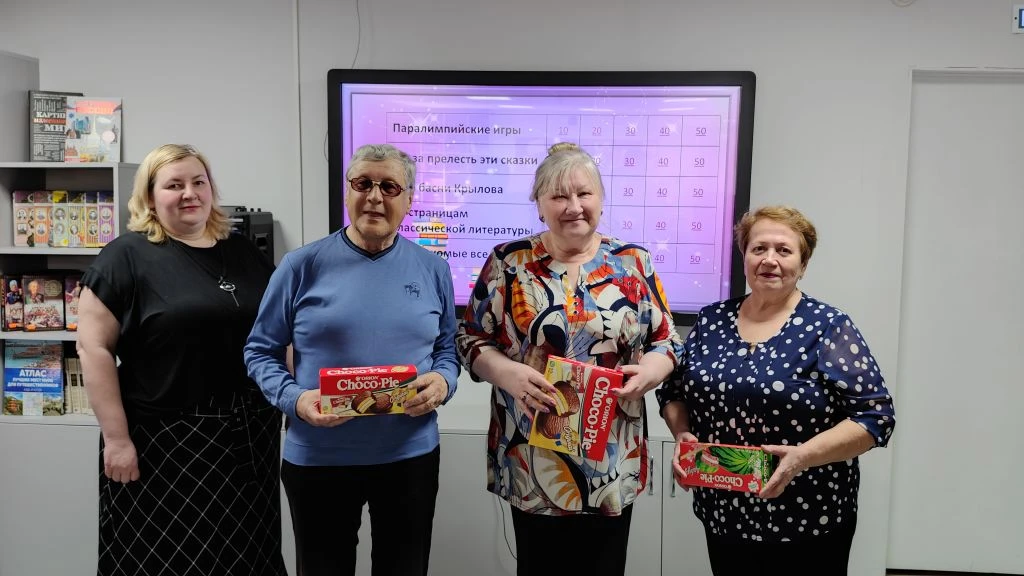Четыре человека стоят и позируют перед экраном; трое держат коробки с печеньем Choco Pie и коробку конфет. На экране позади таблица с темами и баллами, сверху видно «Паралимпийские игры».