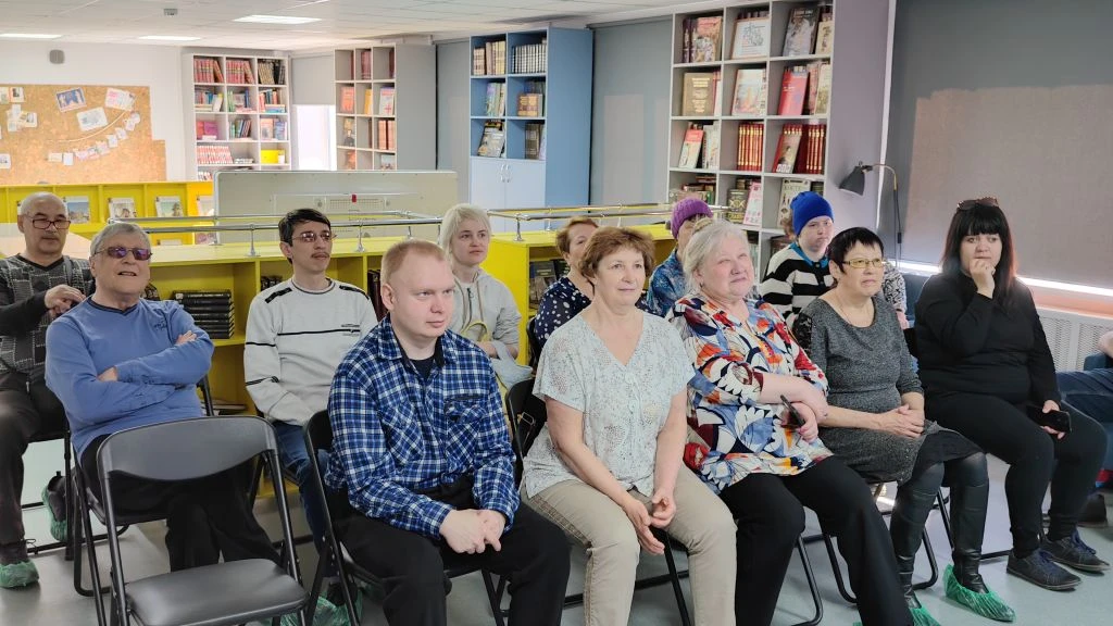 Группа взрослых людей сидит на стульях в библиотеке, все смотрят вперёд. На заднем плане книжные стеллажи.