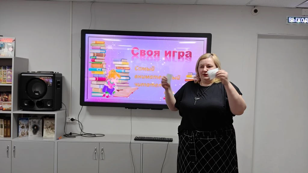 Женщина в комнате стоит перед большим экраном и держит белые листы. На экране слайд «Своя игра» и текст «Самый внимательный читатель». Слева колонка, справа табличка «ВЫХОД».
