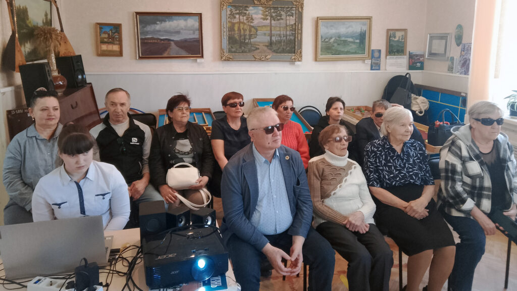 В зале сидят участники мероприятия — мужчины и женщины. Они слушают выступление. Перед ними установлен проектор и ноутбук. Атмосфера спокойная и внимательная.