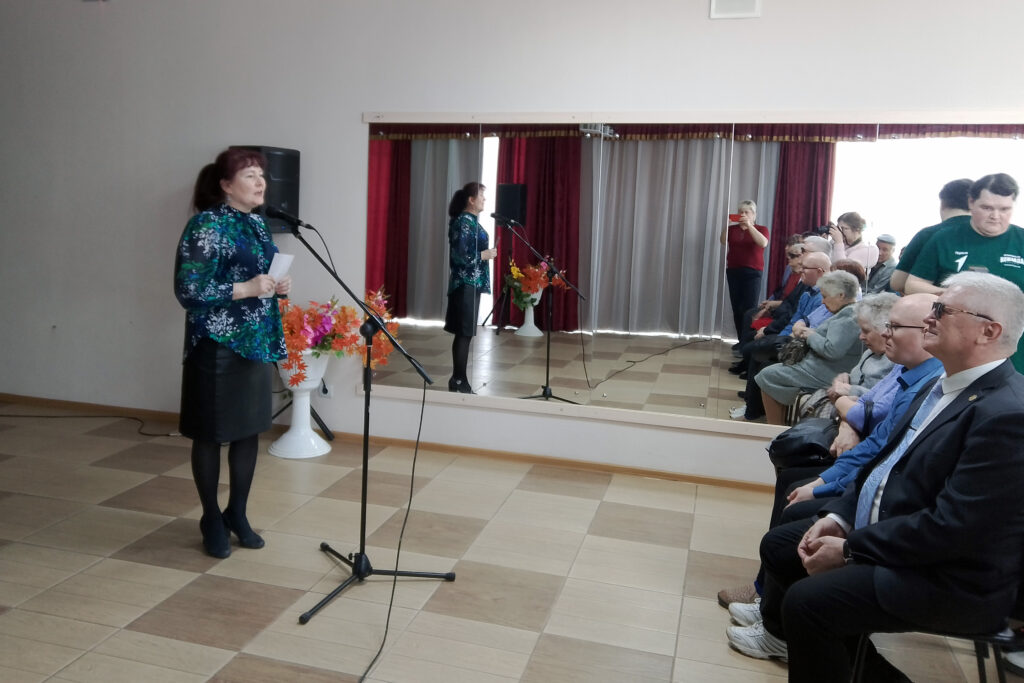 Женщина в зелёно-синей блузке и чёрной юбке стоит у микрофона с листком в руках и выступает перед сидящей аудиторией. Сзади зеркало, бордовые шторы и ваза с яркими цветами; часть людей снимает на телефон.