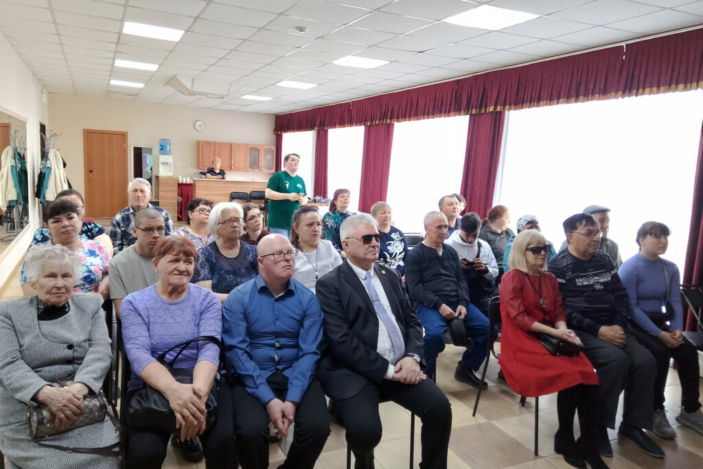 На фото аудитория в небольшом зале: несколько рядов сидящих людей, слушают выступление. На заднем плане большие окна с бордовыми шторами; у стены видны вешалки с одеждой и стойка/буфет, рядом стоит человек в зелёной футболке.