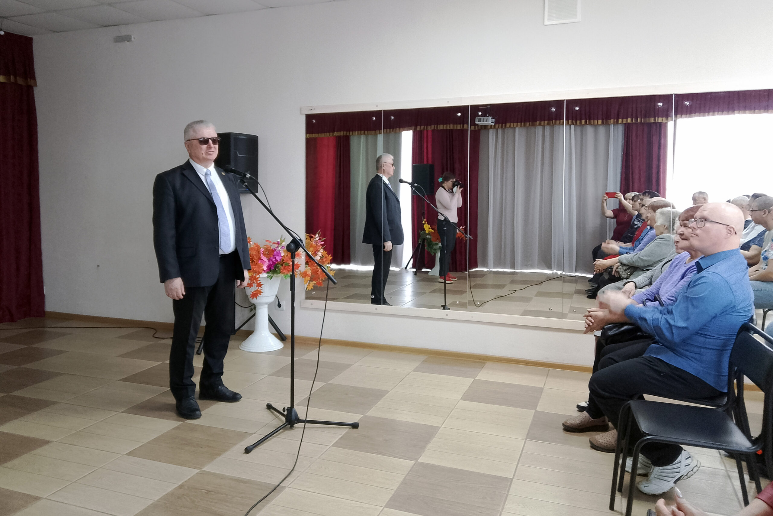 Мужчина в чёрном костюме и тёмных очках стоит у микрофона и выступает перед сидящей аудиторией; часть людей аплодирует. Сзади большое зеркало и бордовые шторы, сбоку колонка и ваза с яркими цветами.