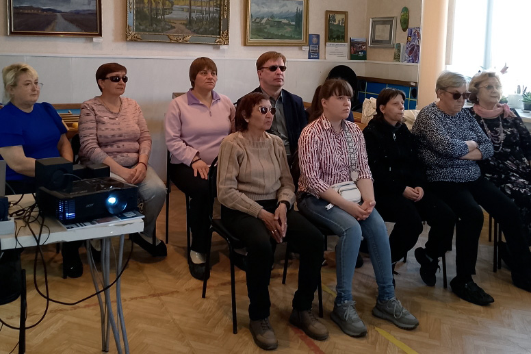 Группа из девяти человек сидит в ряд на стульях в помещении, многие в тёмных очках. Слева на столике стоит включённый проектор. На стене висят картины, справа окна и растения.