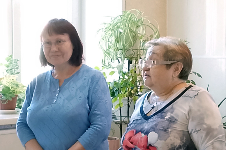 Две женщины в помещении рядом с окнами и комнатными растениями. Слева женщина в голубом свитере и очках, стоит и слегка улыбается. Справа пожилая женщина в очках и светлой кофте с крупным рисунком цветка, повернута влево, как будто говорит.