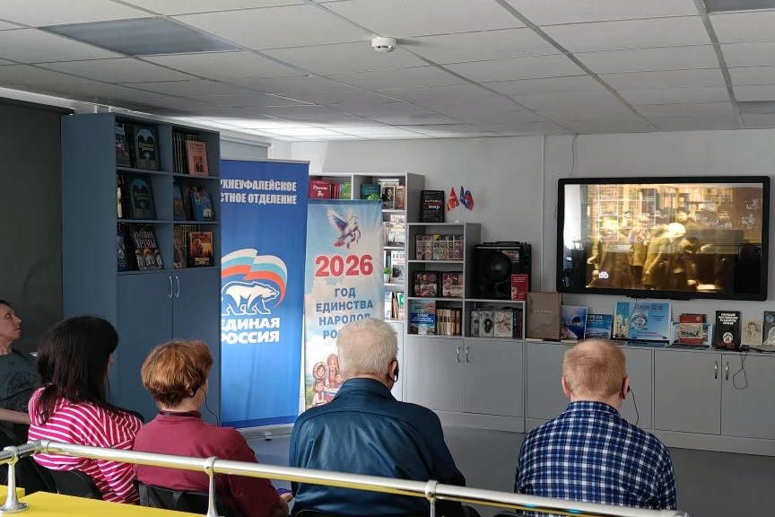 Небольшая аудитория: несколько людей сидят спиной к камере и смотрят видео на большом экране справа. В центре стоят два баннера: «Единая Россия» и «2026 год единства народов России». Вдоль стены шкафы с книгами и выставкой изданий, рядом колонка.