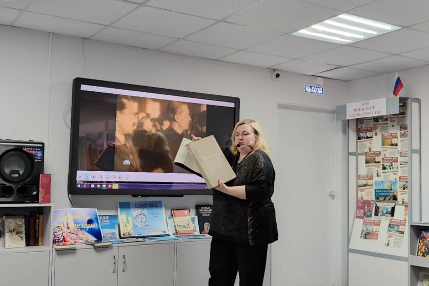 Женщина с гарнитурой выступает и держит раскрытую большую книгу. На стене большой экран с видео, под ним выставка книг; справа дверь с табличкой «ВЫХОД» и стенд с брошюрами «ВОИНСКОЙ СЛАВЫ РОССИИ».