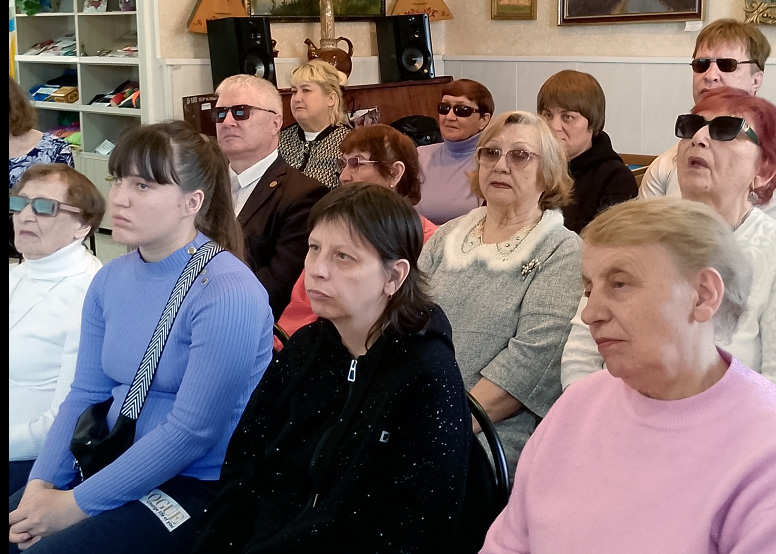 Группа людей сидит в зале на мероприятии. На переднем плане три женщины; позади ещё несколько мужчин и женщин, многие в тёмных очках.