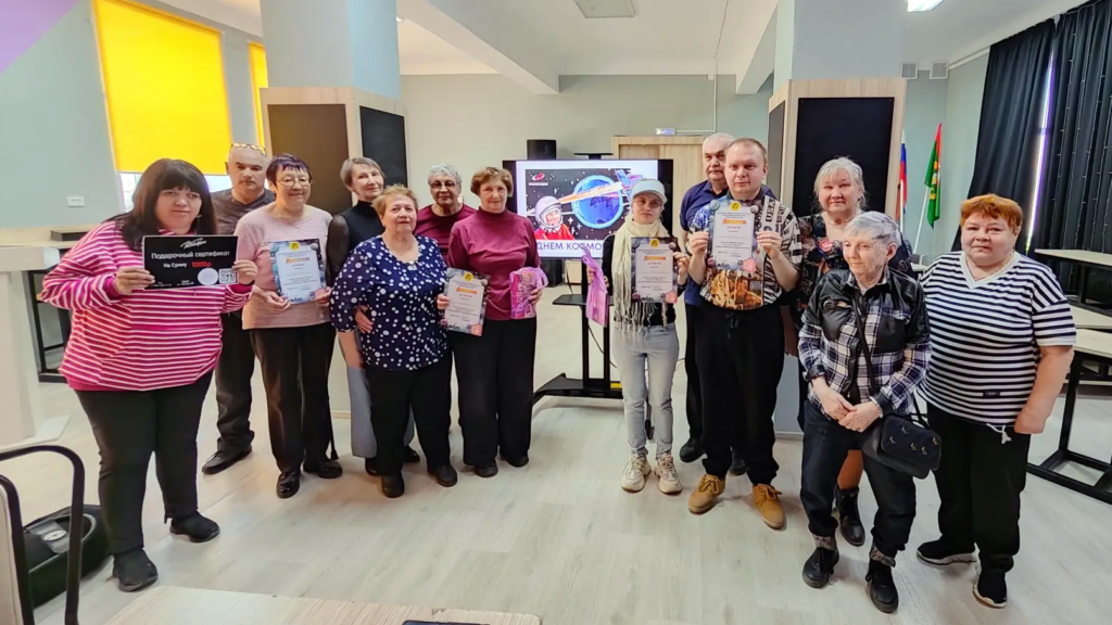 Группа из 14 человек стоит в помещении и позирует с дипломами и подарками на фоне экрана с изображением ко Дню космонавтики. Слева женщина в розовой полосатой кофте держит большой подарочный сертификат на сумму 1000. Остальные в руках держат дипломы и розовые подарочные пакеты. Все выглядят довольными.