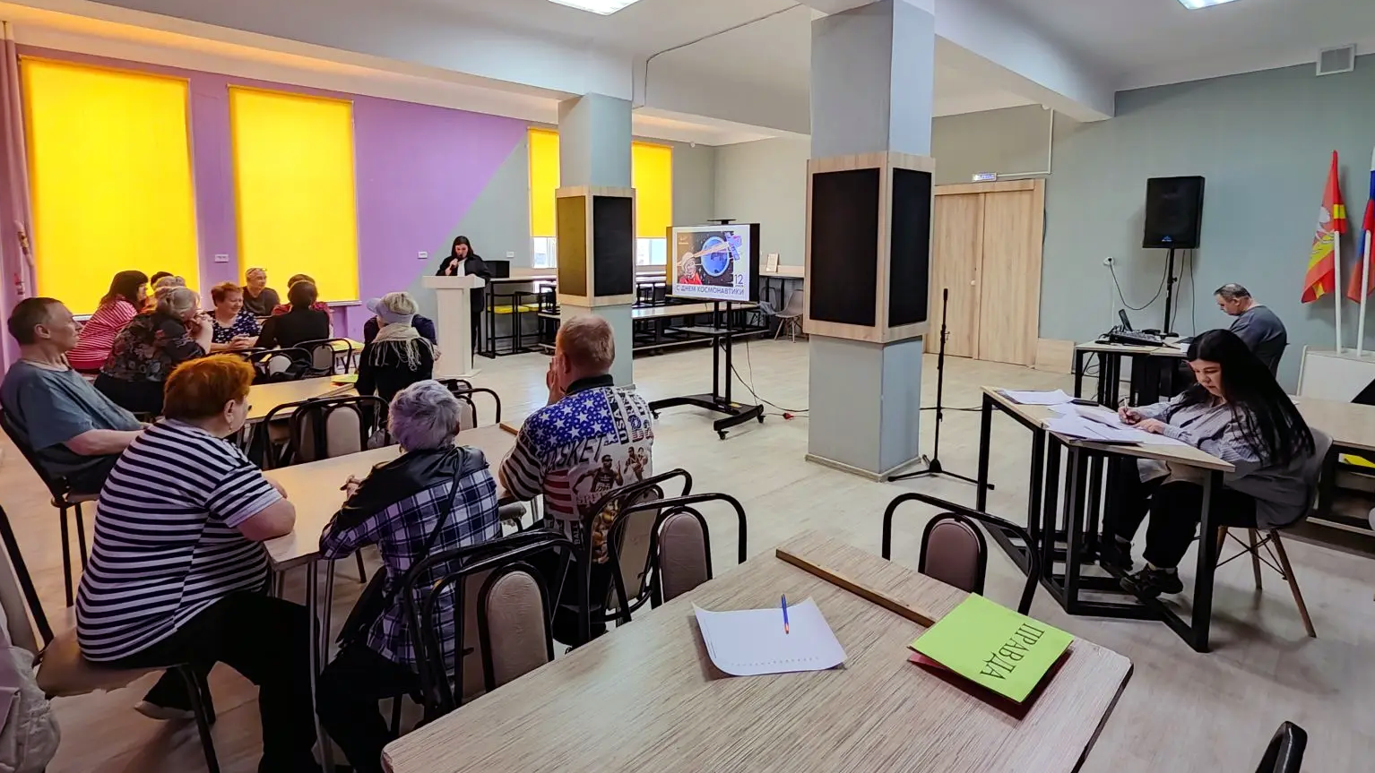 в комнате проходит мероприятие ко Дню космонавтики: люди сидят за столами, впереди женщина у кафедры, в центре экран с надписью «С ДНЕМ КОСМОНАВТИКИ», на ближнем столе папка с надписью «ИГРА».