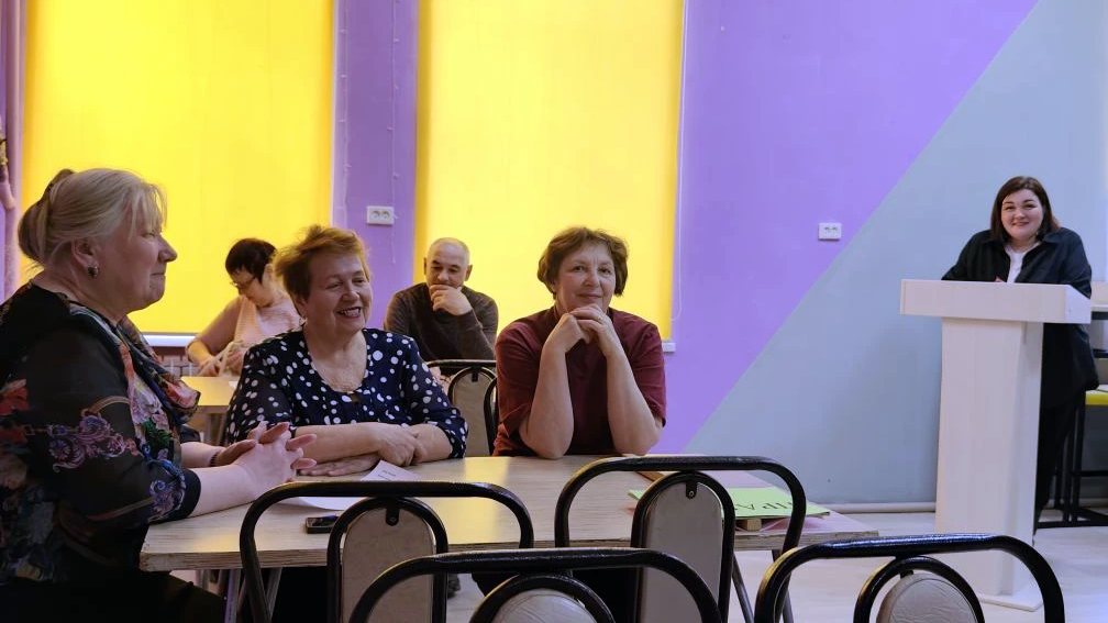 На переднем плане за столом сидят три женщины, улыбаются и общаются. На фоне ещё несколько людей за столами. Справа у кафедры стоит женщина, тоже улыбается. На стенах яркие жёлтые и сиреневые панели.