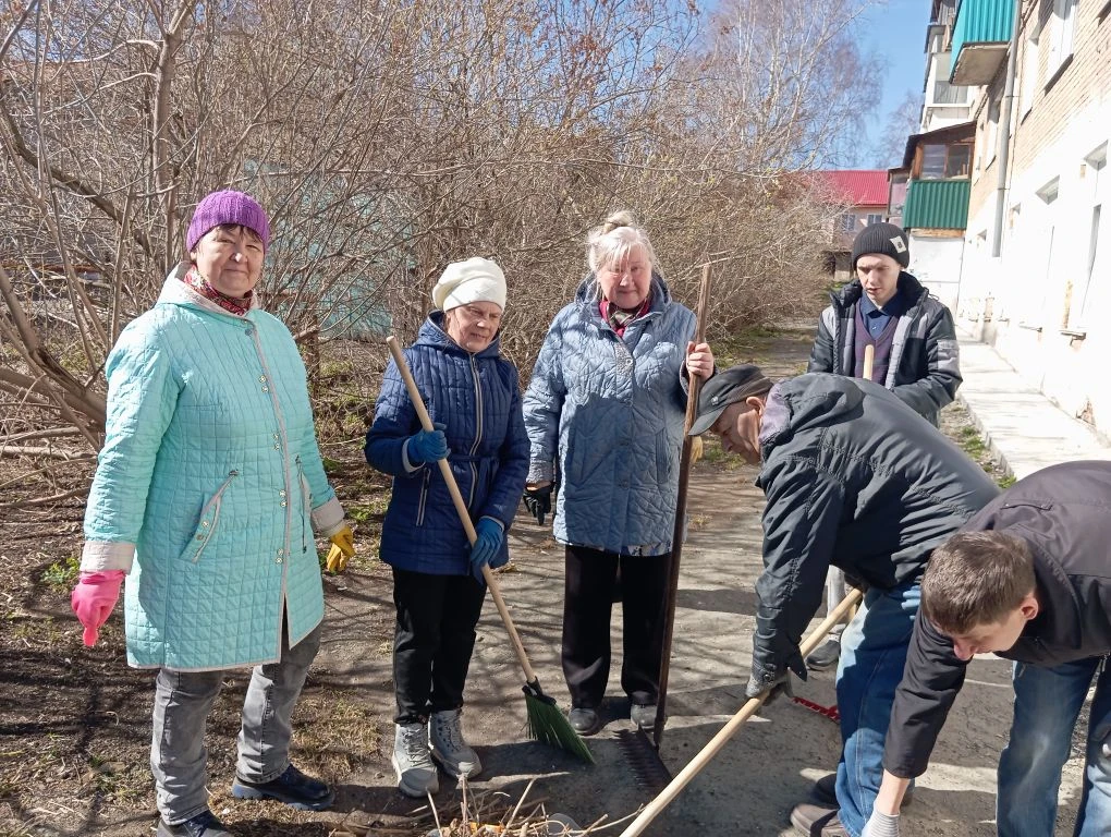 Люди во дворе у дома делают уборку: стоят с граблями и метлой на дорожке в солнечный день.